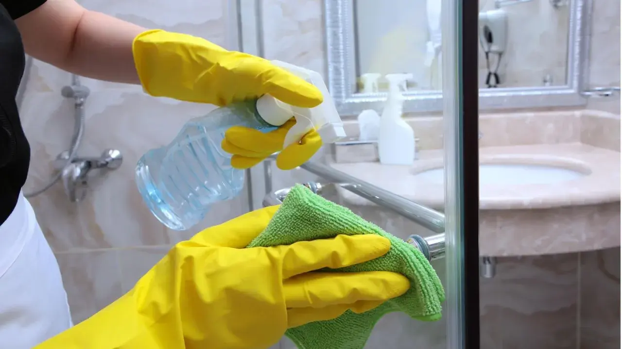 Hände in gelben Handschuhen sprühen Reinigungsmittel auf eine Duschkabine und wischen sie mit einem grünen Tuch ab.