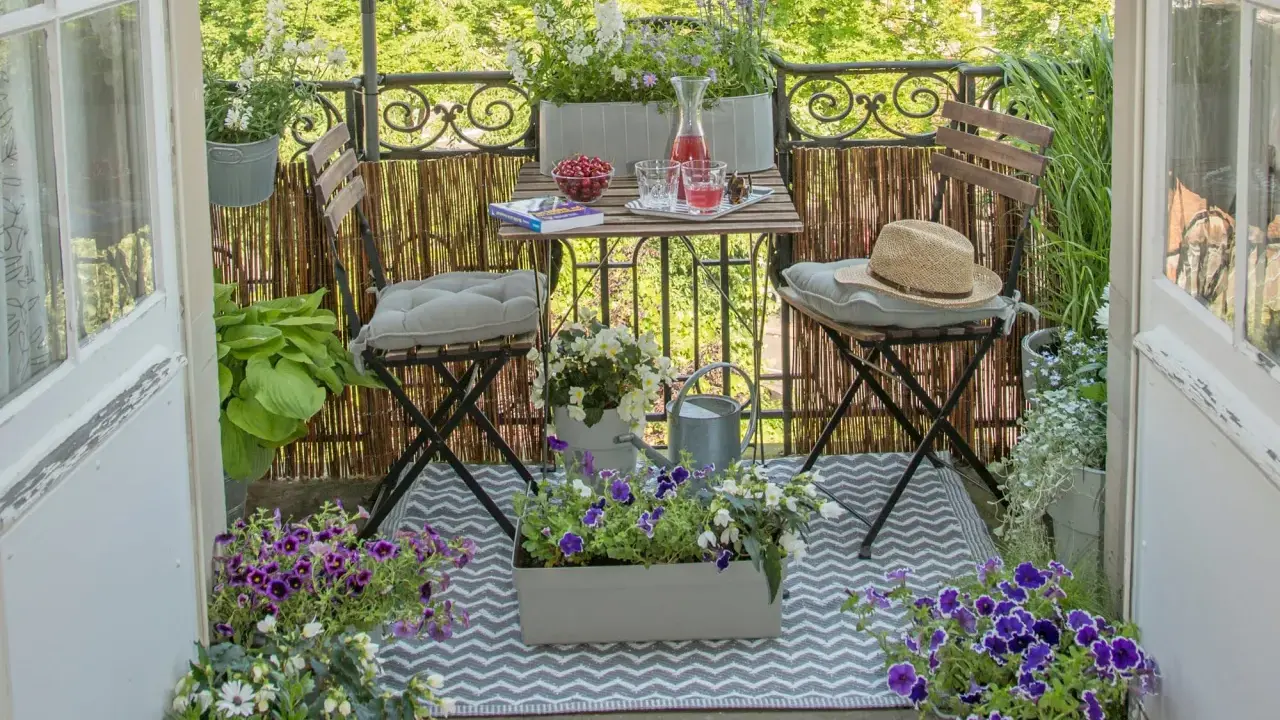 Gemütliche Balkon Ideen: Ein kleiner Tisch mit Getränken und Kirschen, zwei Stühle mit Kissen, Blumen und eine Gießkanne laden zum Verweilen ein.