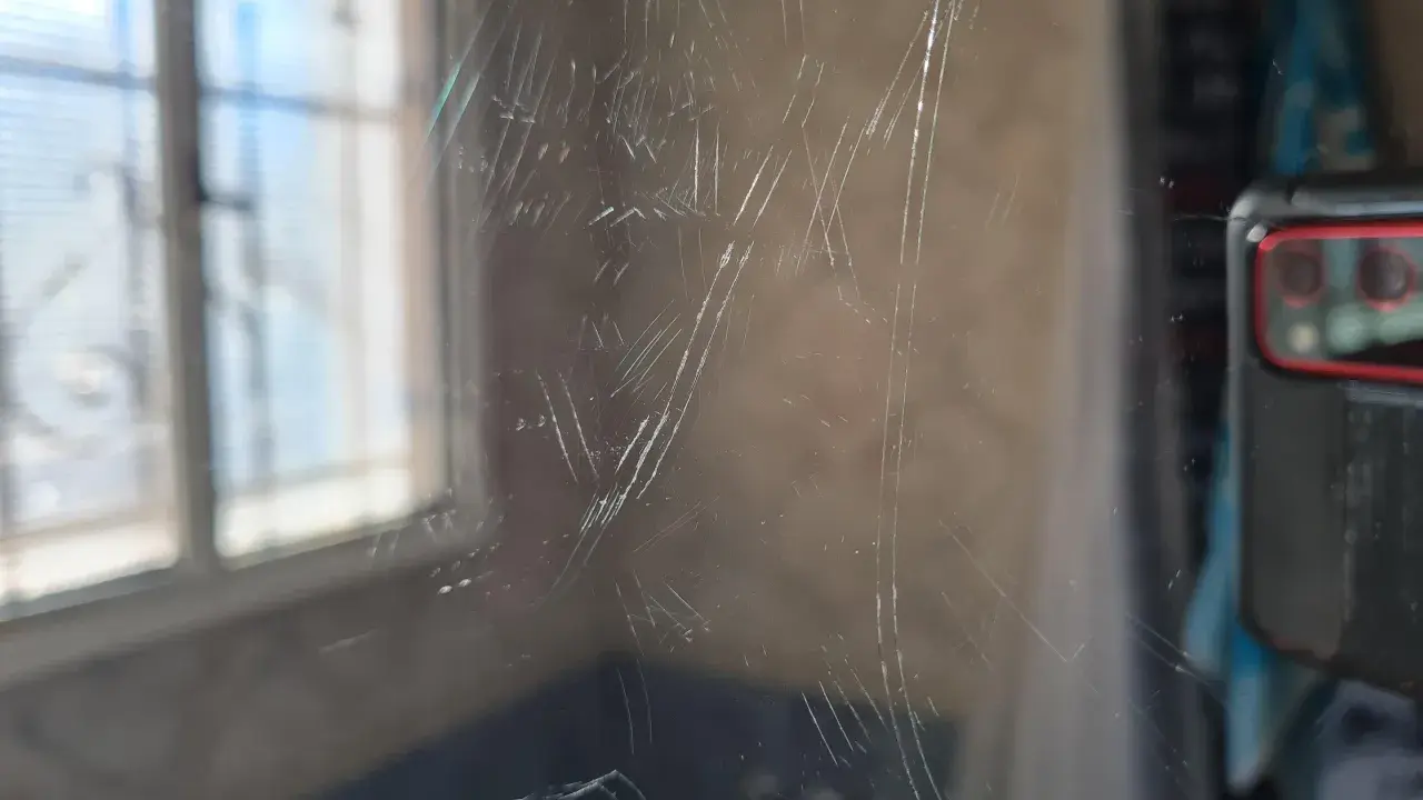 Zahlreiche Kratzer auf einer Glasoberfläche, die auf eine Politur warten. Im Hintergrund ist ein Fenster mit Jalousien zu sehen.