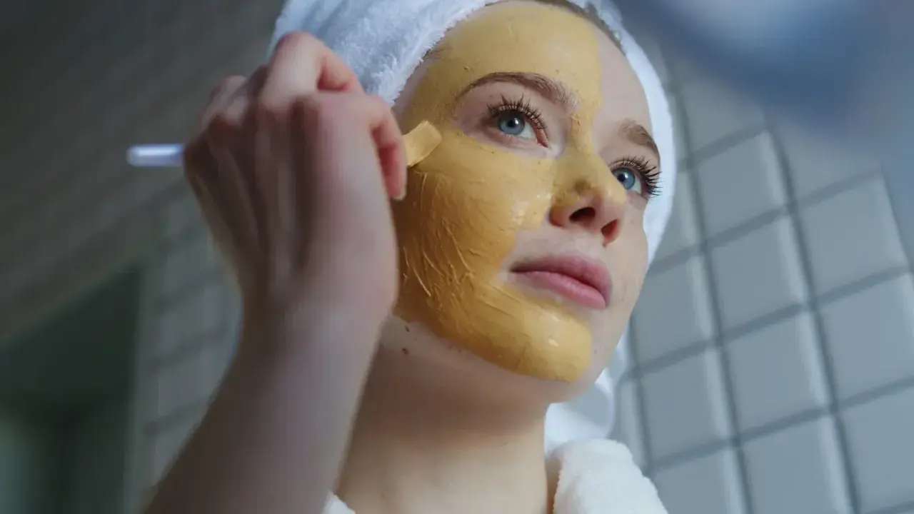 Junge Frau mit Handtuch auf dem Kopf trägt eine Kurkuma-Maske auf. Die Kurkuma Wirkung auf die Haut ist bekannt für ihre entzündungshemmenden Eigenschaften.