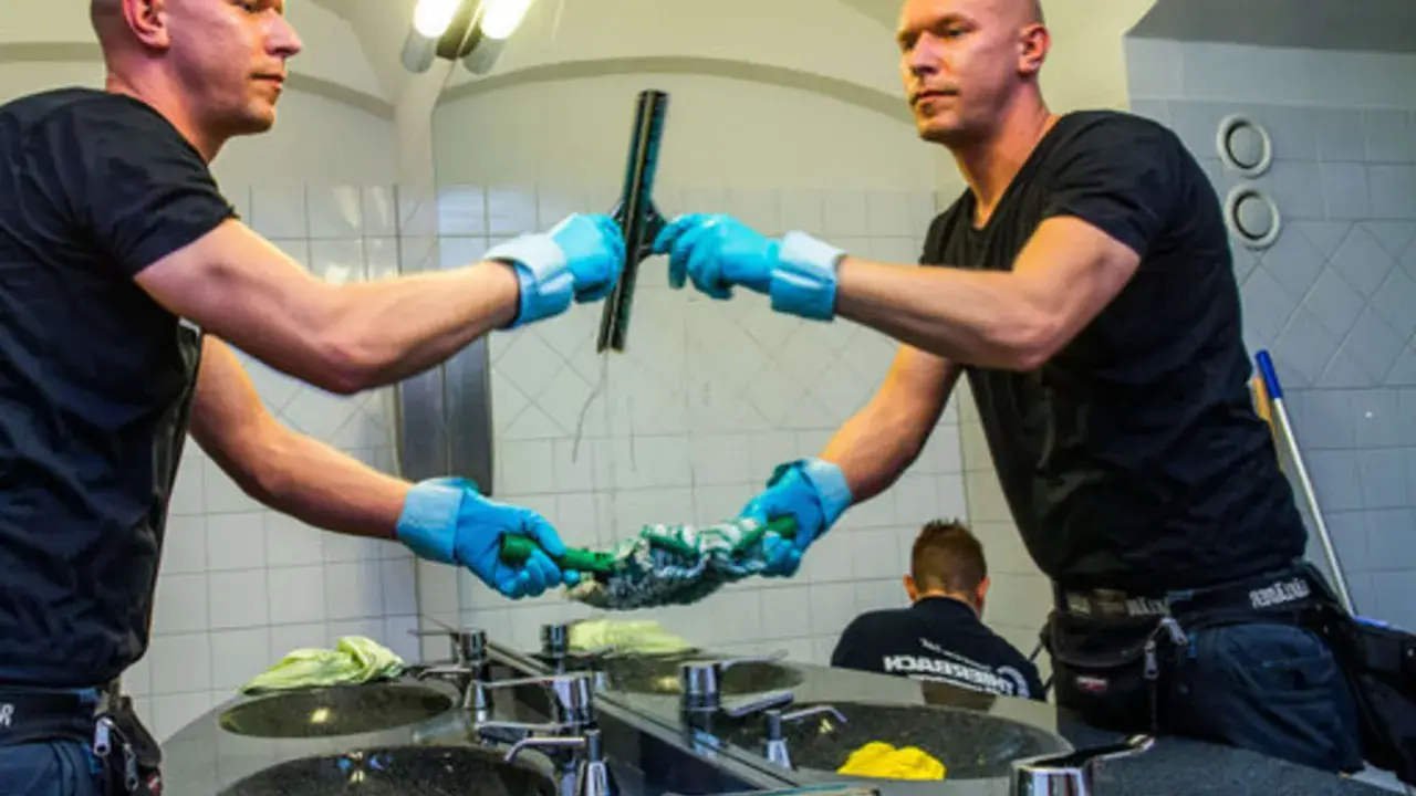 Mann mit blauen Handschuhen putzt mit einem Abzieher den Spiegel.
