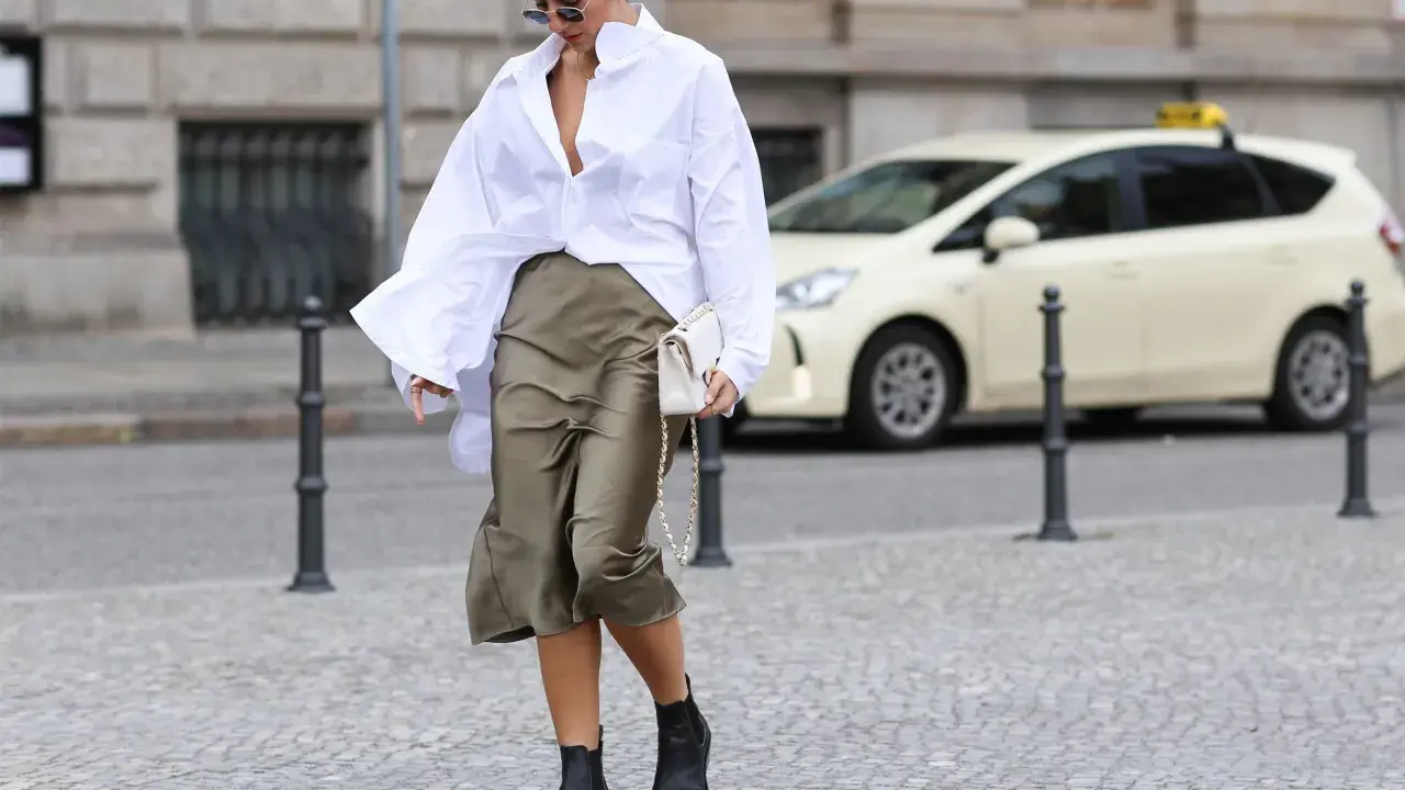 Eine Frau stylt eine weiße Oversize Bluse mit einem Satinrock und schwarzen Stiefeln. Sie trägt eine weiße Tasche.