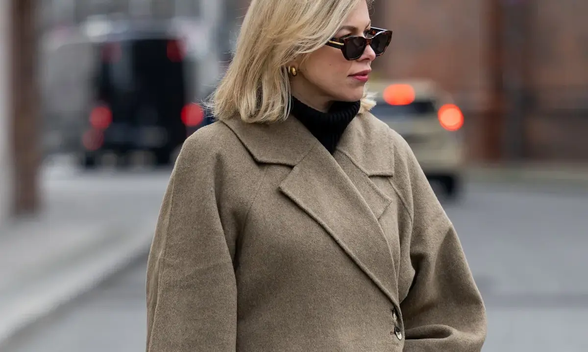 Eine Frau mit blondem Scandi Bob und Sonnenbrille trägt einen beigen Wollmantel und schwarzen Rollkragenpullover.