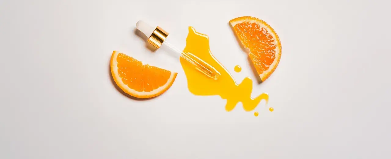 Ein Pipettenfläschchen mit goldenem Detail tropft ein leuchtendes Vitamin C Serum auf eine weiße Fläche, flankiert von zwei saftigen Orangenscheiben. Morgens oder abends für strahlende Haut.