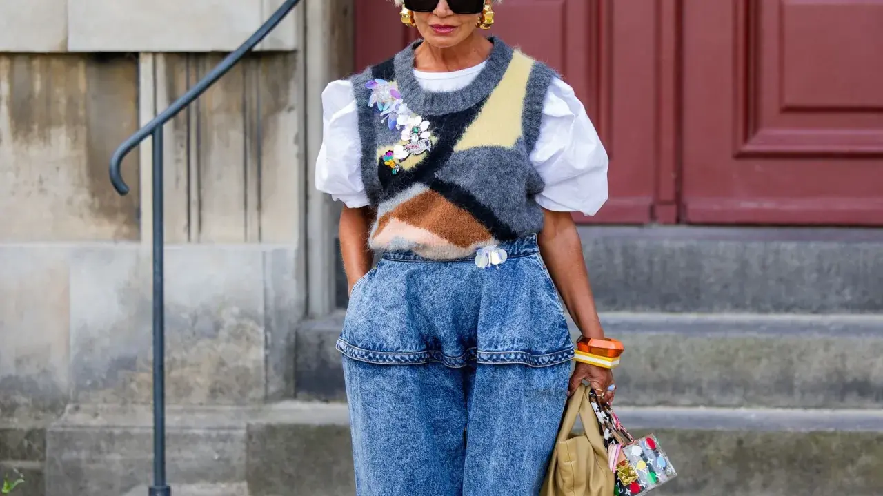 Modische Frau mit weißem Haar, Sonnenbrille und auffälligem Pullunder, der lässig zu Jeans kombiniert wird.