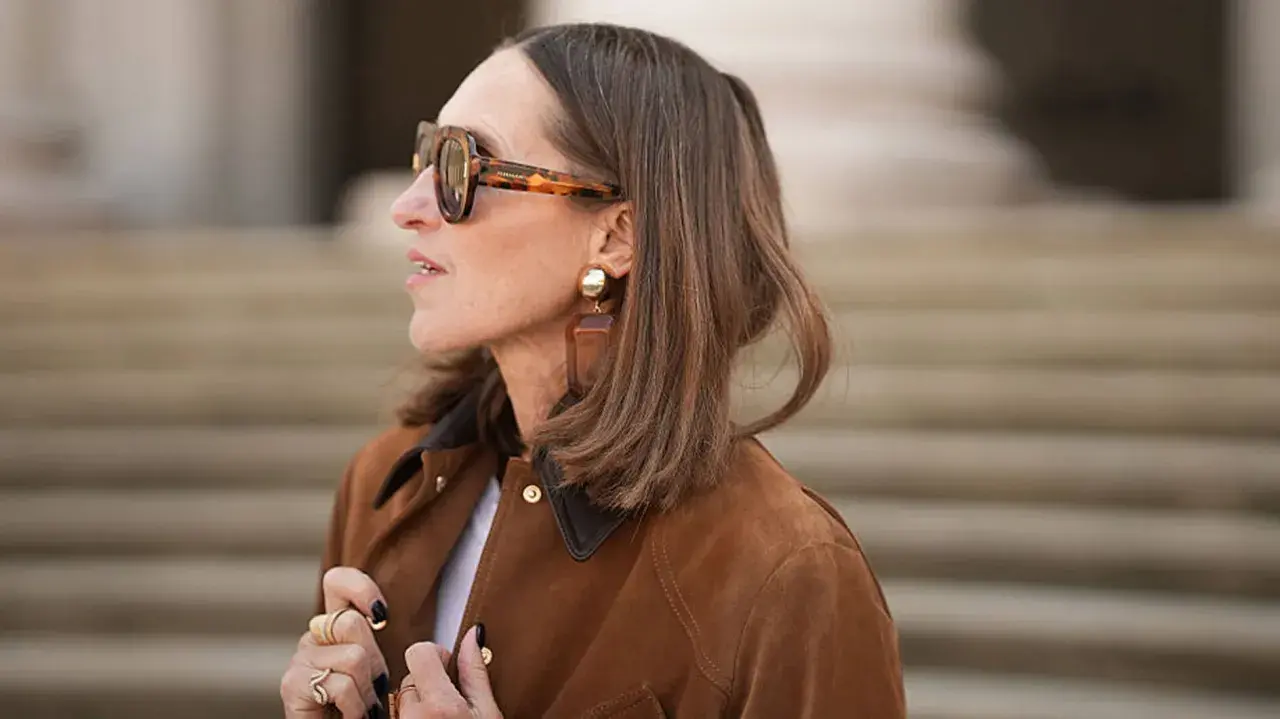 Eine Frau mit braunem Bob und auffälliger Sonnenbrille trägt eine braune Wildlederjacke. Sie erinnert an den Stil von Rodeo Bob.
