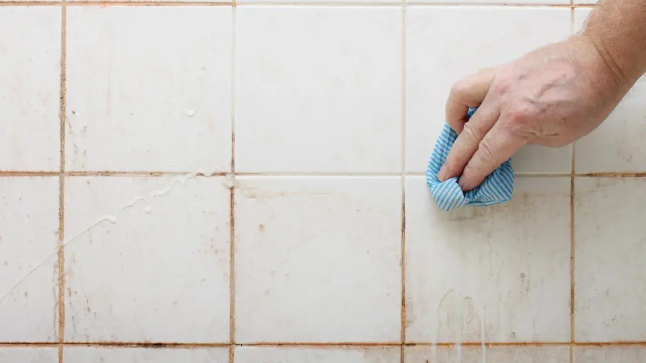 Hand reinigt schmutzige weiße Wandfliesen mit einem blauen Tuch.