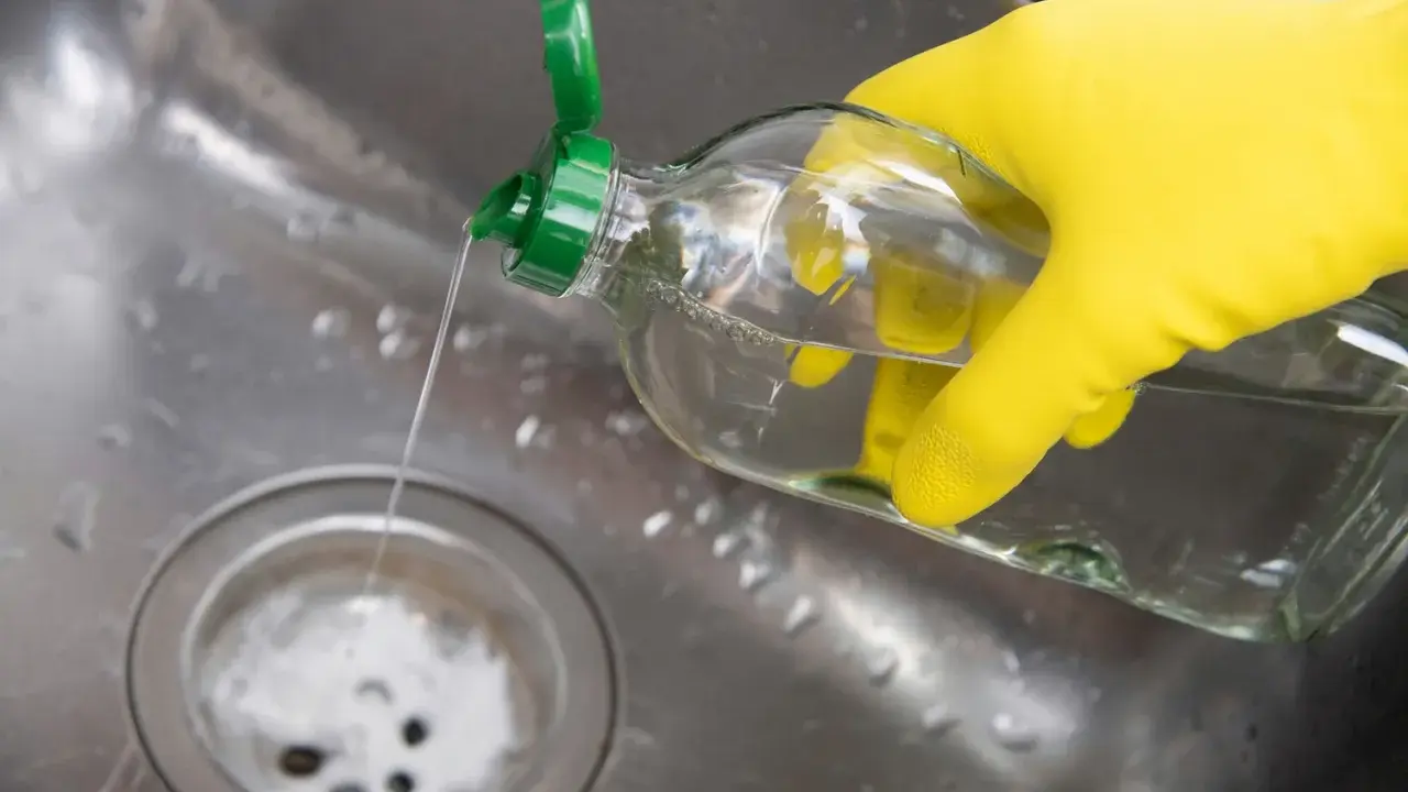 Gelbe Handschuhe halten eine Flasche mit klarem Flüssigkeit, die in ein Spülbecken gegossen wird. Ein Hausmittel Rohrreiniger, der Abfluss wird gereinigt.