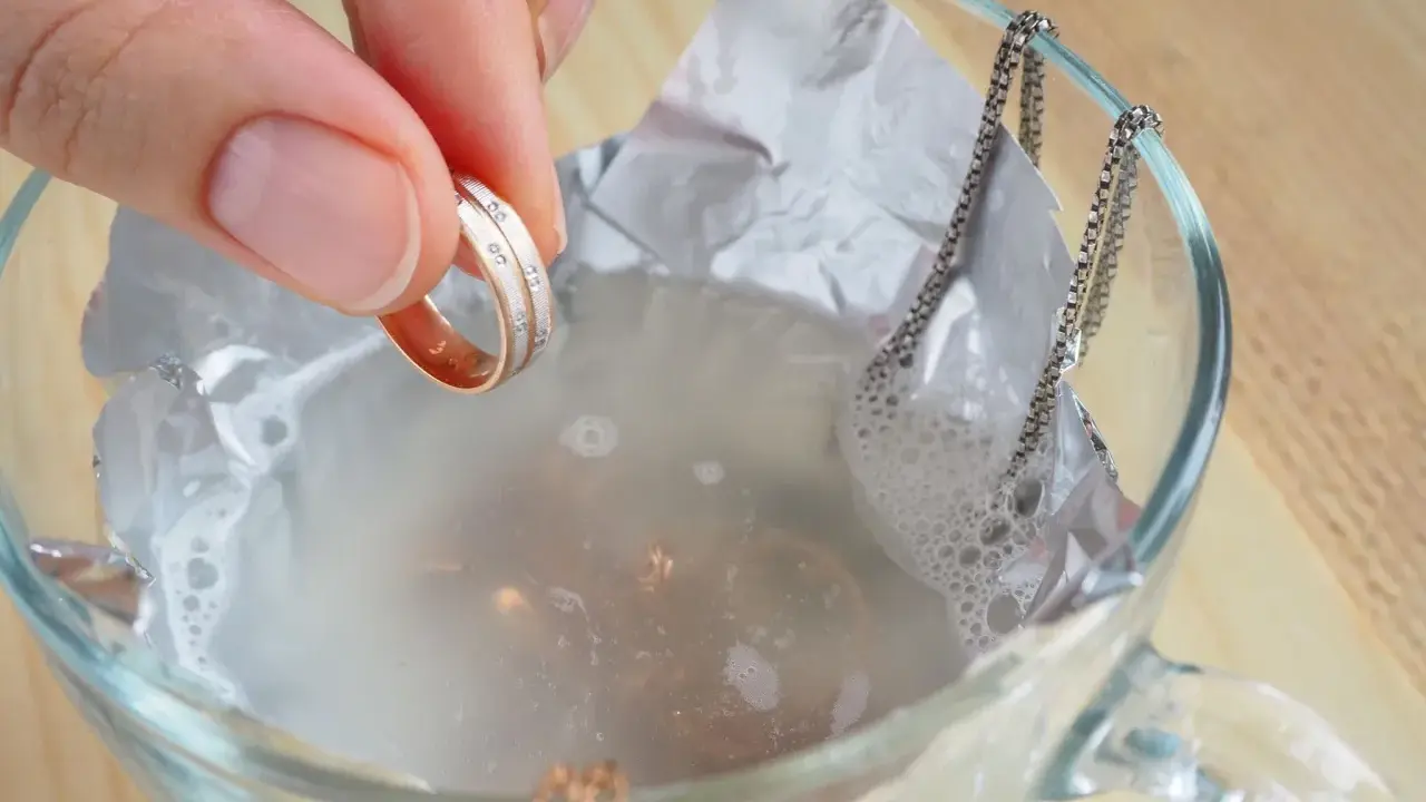 Schmuckreinigung mit Hausmitteln: Eine Hand hält einen Ring, während eine Silberkette in einer Schüssel mit Wasser und Alufolie liegt.