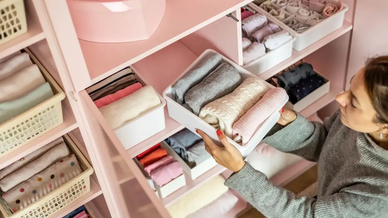 Frau sortiert Kleidung in einem rosa Kleiderschrank. Sie zeigt, wie man den Kleiderschrank organisiert, mit gefalteten Pullovern in weißen Boxen.