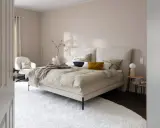 Beige schlafzimmer ideen: einladendes Bett mit beiger Bettwäsche, Akzentkissen und Decke. Weißer Sessel, Nachttische und Teppich schaffen Gemütlichkeit.