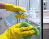 Hände in gelben Handschuhen sprühen Reinigungsmittel auf eine Duschkabine und wischen sie mit einem grünen Tuch ab.