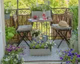 Gemütliche Balkon Ideen: Ein kleiner Tisch mit Getränken und Kirschen, zwei Stühle mit Kissen, Blumen und eine Gießkanne laden zum Verweilen ein.