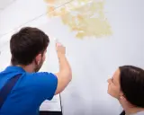 Mann prüft Stockflecken an der Wand, um sie zu entfernen. Eine Frau schaut besorgt zu.