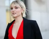 Blonde Frau mit einer stylischen Frisur zwischen Pixie und Bob, trägt einen roten V-Ausschnitt und einen schwarzen Blazer. Goldene Ohrringe setzen Akzente.