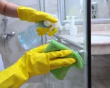 Hände in gelben Handschuhen sprühen Reinigungsmittel auf die Duschkabine, um Kalkflecken am Glas zu entfernen.