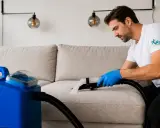 Mann mit blauen Handschuhen reinigt mit einem Sprühextraktionsgerät eine beige Couch.