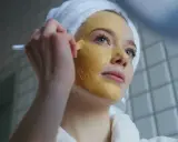 Junge Frau mit Handtuch auf dem Kopf trägt eine Kurkuma-Maske auf. Die Kurkuma Wirkung auf die Haut ist bekannt für ihre entzündungshemmenden Eigenschaften.
