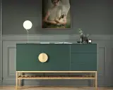 Ein grünes Sideboard mit Holzdetails, dekoriert mit einer Lampe, einem Buch und Vasen. Ein Gemälde mit einem Schwan hängt darüber.