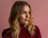 Blonde Frau mit einem **kitty cut**-Haarschnitt, trägt einen roten Rollkragenpullover vor rosafarbenem Hintergrund.