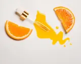 Ein Pipettenfläschchen mit goldenem Detail tropft ein leuchtendes Vitamin C Serum auf eine weiße Fläche, flankiert von zwei saftigen Orangenscheiben. Morgens oder abends für strahlende Haut.