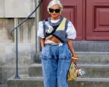 Modische Frau mit weißem Haar, Sonnenbrille und auffälligem Pullunder, der lässig zu Jeans kombiniert wird.
