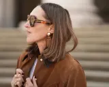 Eine Frau mit braunem Bob und auffälliger Sonnenbrille trägt eine braune Wildlederjacke. Sie erinnert an den Stil von Rodeo Bob.