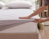 Hände legen ein graues Spannbettlaken ordentlich auf eine Matratze.