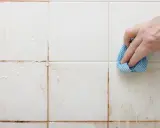 Hand reinigt schmutzige weiße Wandfliesen mit einem blauen Tuch.