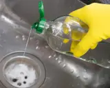 Gelbe Handschuhe halten eine Flasche mit klarem Flüssigkeit, die in ein Spülbecken gegossen wird. Ein Hausmittel Rohrreiniger, der Abfluss wird gereinigt.