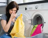 Frau hält sich die Nase zu, weil die Waschmaschine stinkt. Gelbes Handtuch in der Hand, blickt sie angewidert auf die Wäsche.