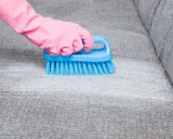 Hand in pink glove uses blue brush to scrub grey fabric, showing how to remove blutflecken.