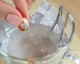 Schmuckreinigung mit Hausmitteln: Eine Hand hält einen Ring, während eine Silberkette in einer Schüssel mit Wasser und Alufolie liegt.