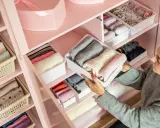 Frau sortiert Kleidung in einem rosa Kleiderschrank. Sie zeigt, wie man den Kleiderschrank organisiert, mit gefalteten Pullovern in weißen Boxen.