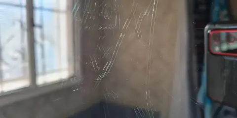 Zahlreiche Kratzer auf einer Glasoberfläche, die auf eine Politur warten. Im Hintergrund ist ein Fenster mit Jalousien zu sehen.