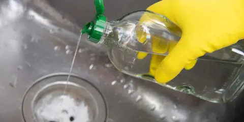 Gelbe Handschuhe halten eine Flasche mit klarem Flüssigkeit, die in ein Spülbecken gegossen wird. Ein Hausmittel Rohrreiniger, der Abfluss wird gereinigt.