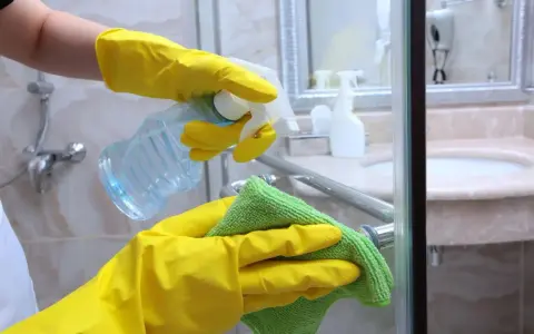 Hände in gelben Handschuhen sprühen Reinigungsmittel auf eine Duschkabine und wischen sie mit einem grünen Tuch ab.