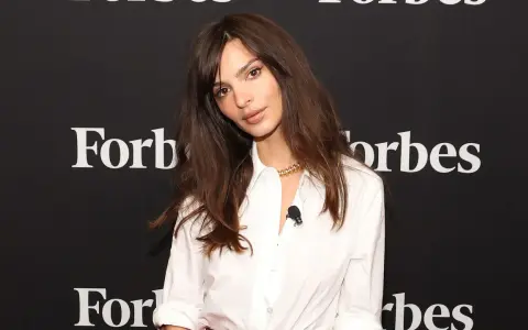 Emily Ratajkowski mit langen, gestuften Haaren und einem weißen Hemd vor einem Forbes-Banner.