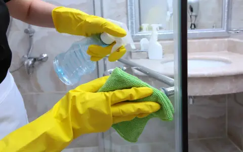 Hände in gelben Handschuhen sprühen Reinigungsmittel auf die Duschkabine, um Kalkflecken am Glas zu entfernen.