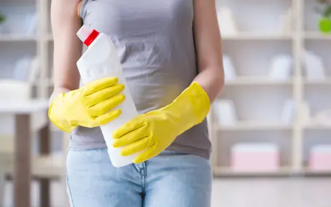 Person in gelben Handschuhen hält Reinigungsmittel. So putzt man Fenster streifenfrei für ein strahlendes Ergebnis.