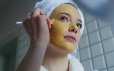 Junge Frau mit Handtuch auf dem Kopf trägt eine Kurkuma-Maske auf. Die Kurkuma Wirkung auf die Haut ist bekannt für ihre entzündungshemmenden Eigenschaften.