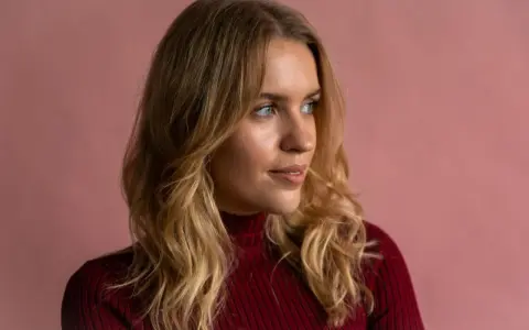 Blonde Frau mit einem **kitty cut**-Haarschnitt, trägt einen roten Rollkragenpullover vor rosafarbenem Hintergrund.