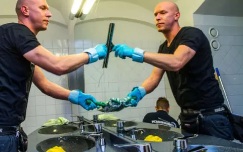 Mann mit blauen Handschuhen putzt mit einem Abzieher den Spiegel.