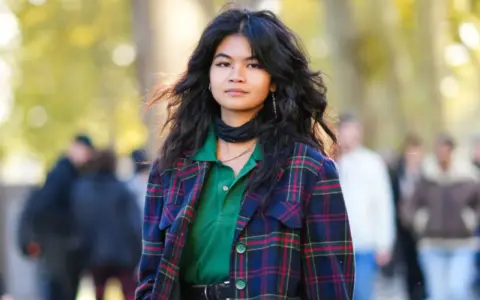 Junge Frau mit voluminösem, flauschigem Haar, trägt karierten Blazer und grünes Poloshirt.