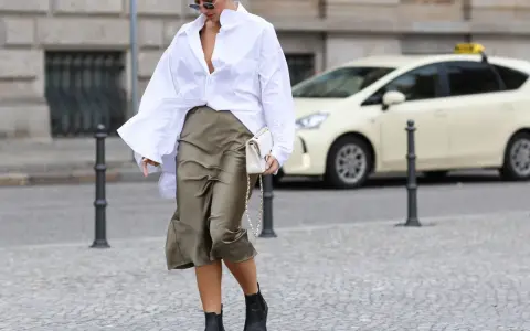 Eine Frau stylt eine weiße Oversize Bluse mit einem Satinrock und schwarzen Stiefeln. Sie trägt eine weiße Tasche.