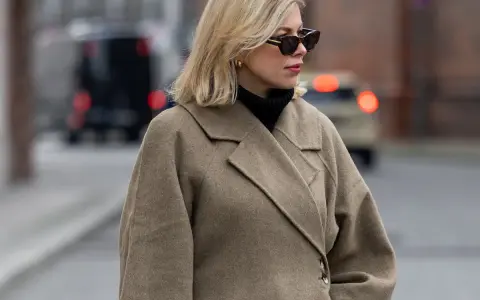 Eine Frau mit blondem Scandi Bob und Sonnenbrille trägt einen beigen Wollmantel und schwarzen Rollkragenpullover.