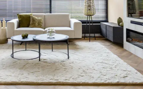 Ein heller, flauschiger Teppich liegt vor einem Sofa und einem Kamin. So wird der Teppich wieder flauschig gemacht.