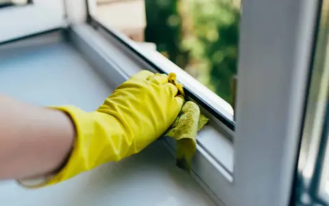 Gelbe Handschuhe reinigen den Kunststoff Fensterrahmen mit einem Tuch.