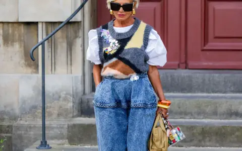 Modische Frau mit weißem Haar, Sonnenbrille und auffälligem Pullunder, der lässig zu Jeans kombiniert wird.