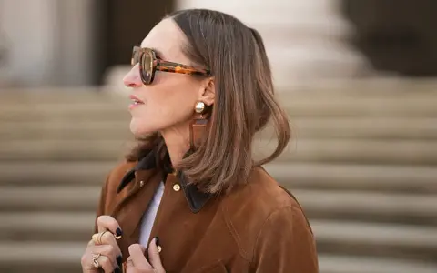 Eine Frau mit braunem Bob und auffälliger Sonnenbrille trägt eine braune Wildlederjacke. Sie erinnert an den Stil von Rodeo Bob.